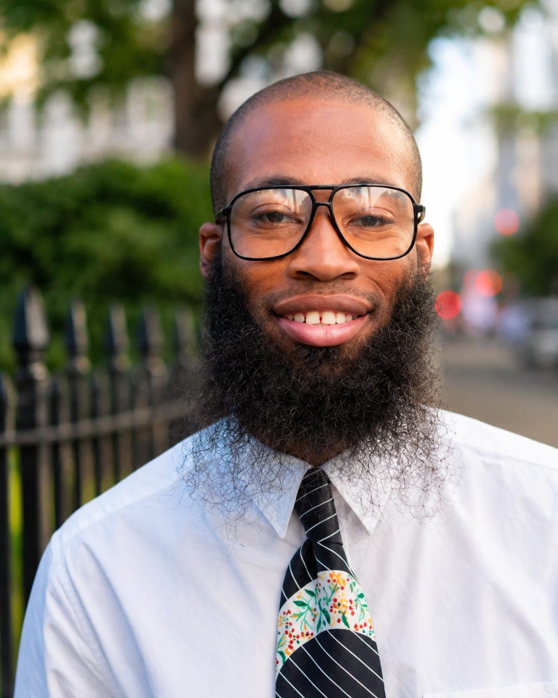 Smiling Black man with a long beard and large glasses wearing a floral tie.