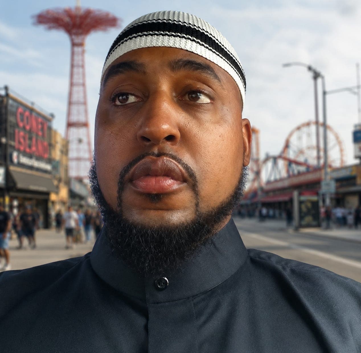 Bearded man in a striped kufi looks upward with Coney Island in the background.