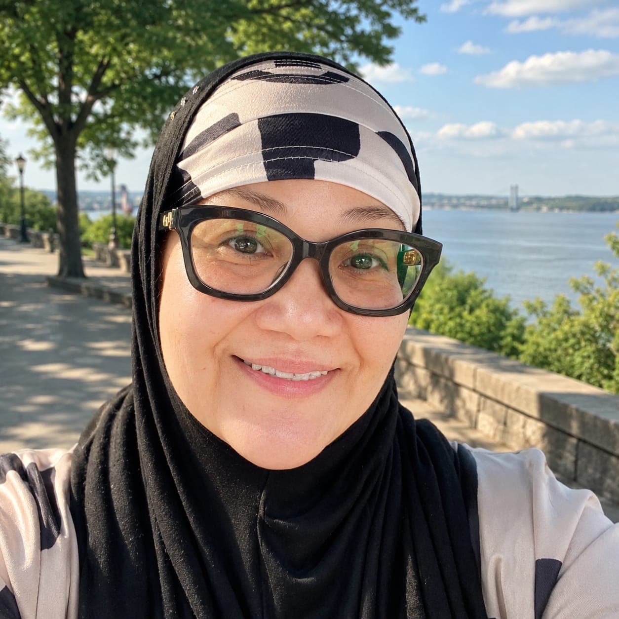 Smiling woman in a black hijab and glasses with a river and bridge background.