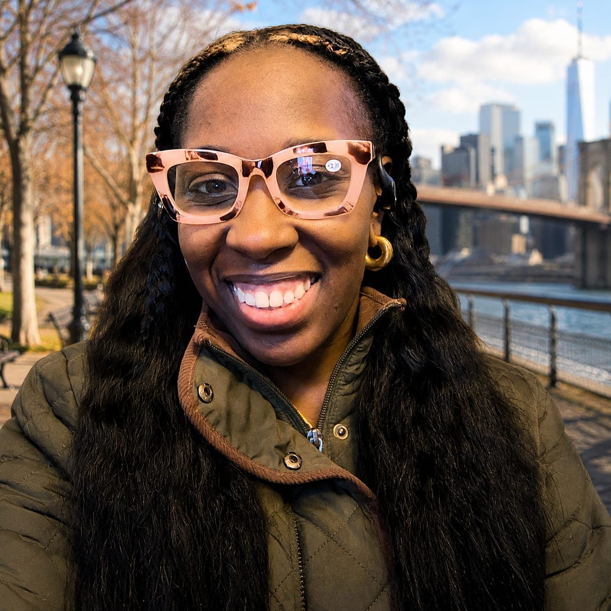 Smiling woman with long braids and pink glasses, with a city skyline and bridge.