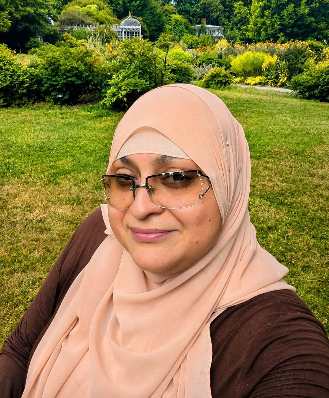 Smiling woman wearing a peach hijab and tinted glasses in a lush green garden.