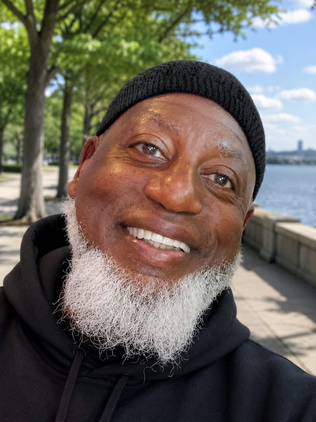 Smiling Black man with a white beard and black beanie, outdoors by the water.